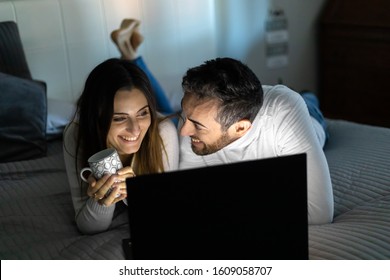 Portrait of a cheerful young couple using laptop computer while laying on bed at night time - Powered by Shutterstock