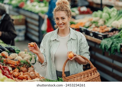 Portrait of a a cheerful young female customer standing near market stand with wicker shopping basket and choosing onions at green market while smiling at camera. Shopper woman buying vegetables. - Powered by Shutterstock