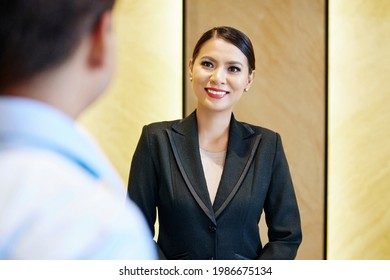Portrait Of Cheerful Young Female Hotel Manager Greeting Guest At Reception Table