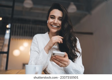 Portrait of cheerful young female with long dark hair in casual clothes enjoying music through TWS earbuds while browsing mobile phone sitting at table - Powered by Shutterstock