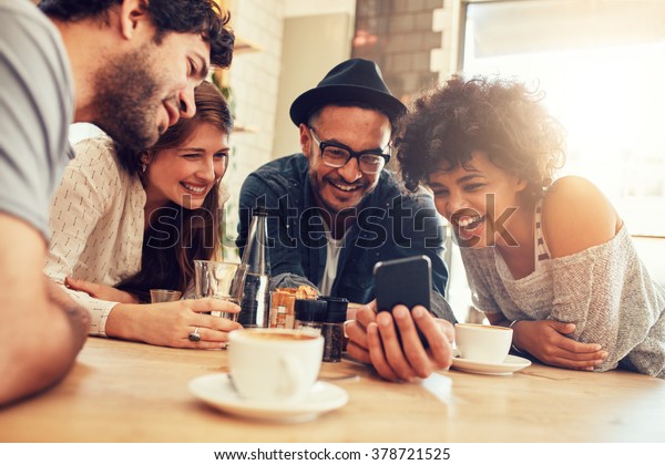 Portrait of cheerful young friends looking\
at smart phone while sitting in cafe. Mixed race people sitting at\
a table in restaurant using mobile\
phone.