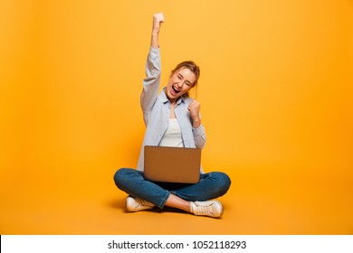 Portrait Of A Cheerful Young Girl With Braces Sitting On A Floor With A Laptop Computer And Celebrating Success Isolated Over Yellow Background