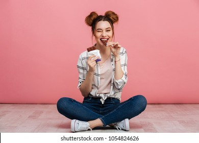 Portrait of a cheerful young girl eating chocolate bar while sitting on a floor with legs crossed isolated over pink background