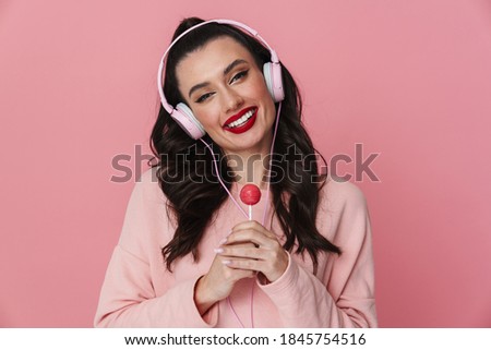 Portrait of a cheerful young girl standing isolated over pink background, wearing headphones, holding lollipop