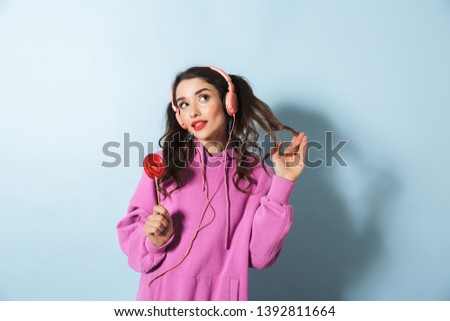 Portrait of a cheerful young girl wearing hoodie standing isolated over blue background, wearing headphones, holding lollipop