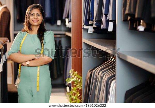Portrait of cheerful young\
Indian shop assistant standing in store with arms folded and\
looking at camera