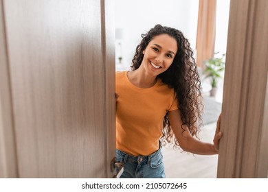 Portrait of cheerful young lady standing in doorway of new modern home, receiving and greeting visitor, happy smiling curly lady holding door looking out of slightly open ajar door