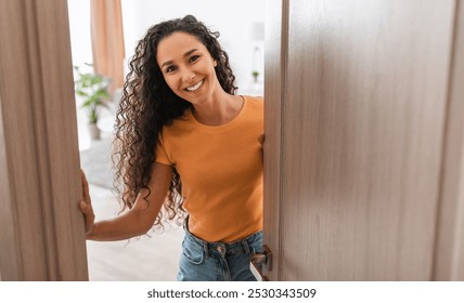 Retrato de la señora joven alegre que se coloca en la puerta del nuevo hogar moderno, recibiendo y saludando al visitante, señora rizada sonriente feliz que sostiene la puerta que mira fuera de la puerta entreabierta ligeramente