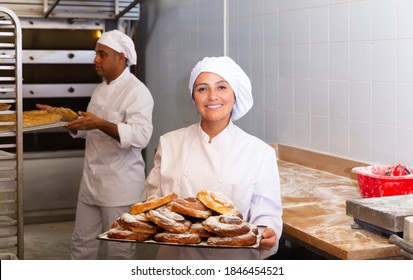 Portrait of cheerful young Latina working in bakery, standing with tray full of sweet traditional Spanish ensaimadas