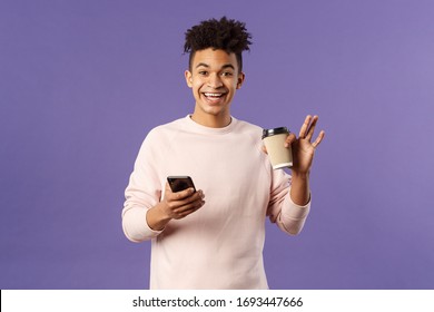 Portrait Of Cheerful Young Male Student With Cup Of Coffee And Mobile Phone, Waving Hand To Say Hi, Smiling Friendly As Greeting Person, Meet Classmate Near University Campus, Purple Background