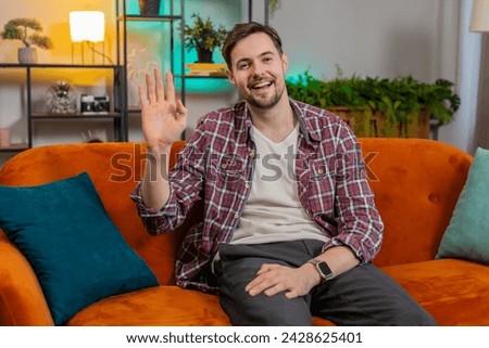 Portrait of cheerful young man in plaid shirt smiling at camera and waving gesturing hello, hi, greeting or goodbye. Happy Caucasian guy making video webcam conference call with friend at home room