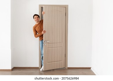 Portrait of cheerful young man standing in doorway of modern apartment, millennial male homeowner holding door looking out and smiling, greeting visitor, full body length - Shutterstock ID 2075626984