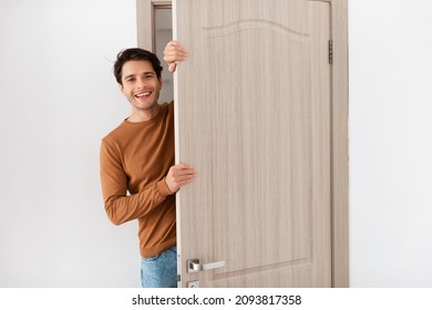 Portrait of cheerful young man standing in doorway of modern apartment, millennial male homeowner holding slightly open ajar door looking out and smiling, greeting visitor, free copy space