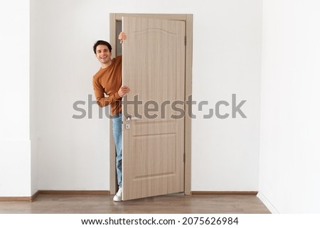Portrait of cheerful young man standing in doorway of modern apartment, millennial male homeowner holding door looking out and smiling, greeting visitor, full body length