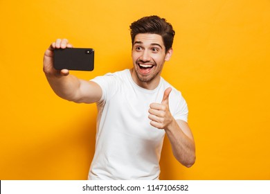 Portrait of a cheerful young man taking a sefie with mobile phone isolated over yellow background, showing thumbs up