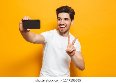 Portrait of a cheerful young man taking a sefie with mobile phone isolated over yellow background