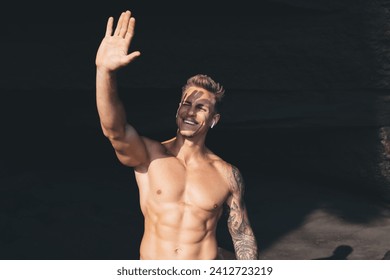 Portrait of cheerful young muscular male with naked torso raising arm and blocking sunlight ray from face with closed eyes while standing against black rock background on sunny day