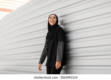 Portrait Of Cheerful Young Muslim Lady Posing In Modest Sportswear While Training Outdoors, Happy Millennial Middle Eastern Lady In Hijab Standing Near Urban Wall, Enjoying Outside Workout