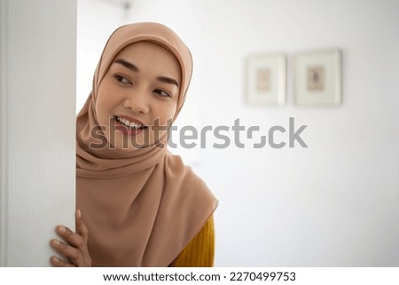 Portrait of cheerful young Muslim woman standing in doorway of modern apartment, millennial female homeowner holding slightly open ajar door looking out and smiling, greeting visitor.