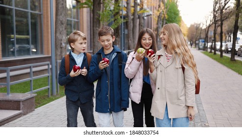 Portrait Of Cheerful Young School Pupils Walking Outdoor And Talking While Eating Apples. Caucasian Boys And Girls College Students With Backpacks Having Snack After School On Street. Lunch Concept