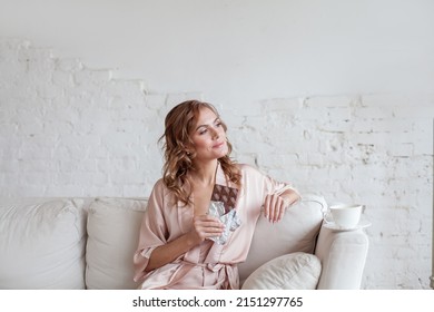 Portrait Of A Cheerful Young Woman Eating Chocolate Bar While Sitting On Sofa And Resting At Home