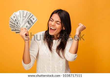 Portrait of a cheerful young woman holding money banknotes and celebrating isolated over yellow background