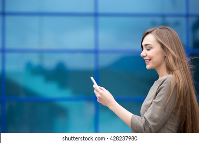 Portrait Of Cheerful Young Woman Sending Message On Smartphone Outdoors. Happy Beautiful Caucasian Woman Using Mobile Phone, Making Call, Looking At Screen On The Street In Summer. Copy Space