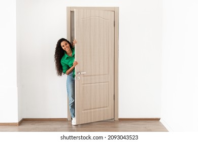 Portrait of cheerful young woman standing in doorway of modern apartment, millennial female homeowner holding slightly open ajar door looking out and smiling, greeting visitor, full body length - Shutterstock ID 2093043523