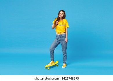 Portrait of cheerful young woman in vivid casual clothes looking camera and standing with yellow skateboard isolated on blue wall background in studio. People lifestyle concept. Mock up copy space