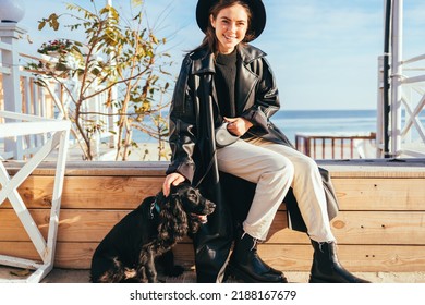 Portrait Cheerful Young Woman Wearing Coat And Hat Petting Her Dog While Sitting On Bench Near Sea On Sunny Day