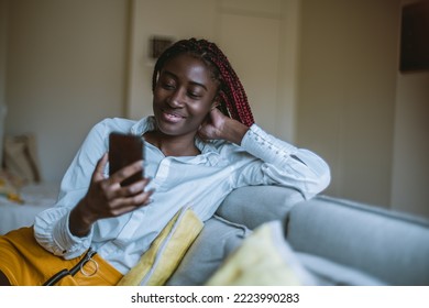 A Portrait Of A Cheerful Youthful Black Female Sitting In Her Room On The Grey Sofa And Having A Video Conversation Via The Smartphone; Young Black Woman Having A Videocall Using Her Cellphone