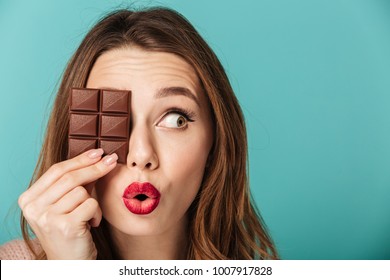 Portrait of a cheery brown haired woman with bright makeup holding chocolate bar at her face isolated over blue background