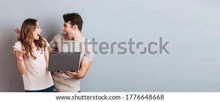 Portrait of a cheery happy couple holding laptop computer while standing and celebrating isolated over gray wall background