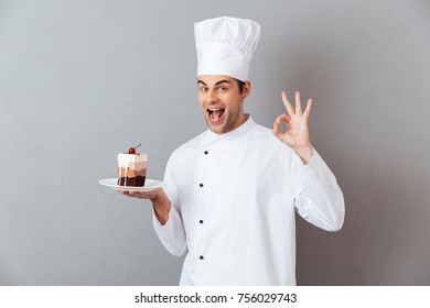 Retrato de un alegre y alegre chef vestido con uniforme de placa sujetador con trozo de pastel y mostrando gesto bueno aislado sobre fondo gris