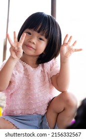 Portrait Of Child 4-5 Year Old. Cute Asian Girl Smiling Sweetly Looking At Camera. Kid Holds Up Four Fingers On One Side Of Three Fingers To Pose For Photo. Vertical Image.