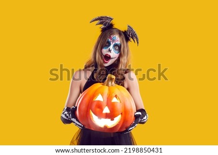 Portrait of child in creative Halloween costume. Girl with skull makeup standing isolated on yellow background, holding Jack o lantern pumpkin and looking at camera with surprised face expression