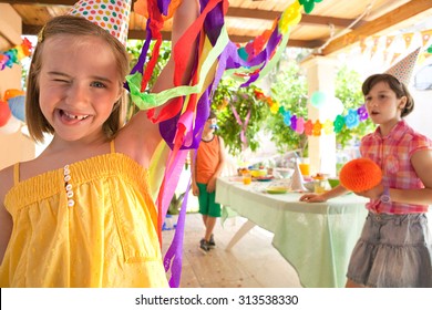 Portrait Of Child Girl With Fun Expression Sticking Her Tongue Out, Being Playful With Friends In A Colorful Summer Birthday Party In A Home Garden, Outdoors. Kids Having Fun Together, Home Exterior.