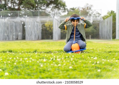 Portrait Of Child With Pogo Stick At Playground. Kid At Park Playing With Plastic Toy. Green Grass, Trees And Nets In Background. Child Is Squatting, Sitting On Knees.