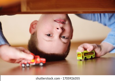 Portrait Of A Child Of A Preschool Boy Upside Down With A Toy Car And A Bus In His Hands. View From The Floor From Under The Bed.