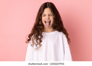 Portrait of childish carefree little girl wearing white T-shirt showing out tongue and closing eyes with naughty disobedient grimace, making face. Indoor studio shot isolated on pink background.