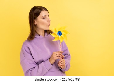 Portrait Of Childish Woman Playing Blowing Yellow Windmill, Having Fun With Paper Toy, Playing With Origami Hand Mill On Stick, Wearing Purple Hoodie. Indoor Studio Shot Isolated On Yellow Background.