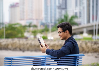 Portrait Of Chinese Businessman Sitting On Bench In Panama City With Skyline In Background, Reading Emails On Tablet Computer