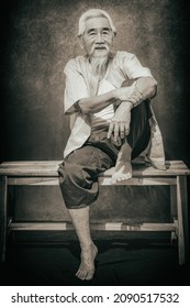 Portrait Of Chinese Old Man With White Hair And Beard In Tradional Chinese Clothing Sitting On Wooden Bench On Studio Backdrop Background In Vintage Tone