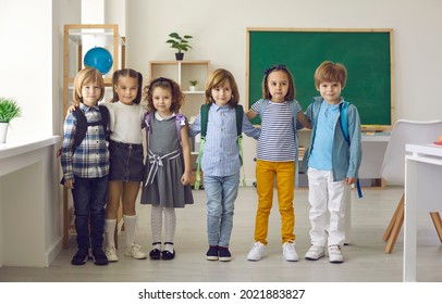 Portrait In The Classroom Of A Small Group Of Elementary School Or Kindergarten Students. Kids In Casual Clothes And With Backpacks On Their Shoulders Are Standing Hugging And Looking At The Camera.