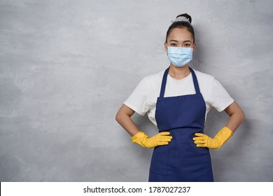 Portrait Of A Cleaning Lady Wearing Yellow Rubber Gloves And Medical Protective Face Mask Keeping Arms On Hips, Looking At Camera While Standing Against Grey Wall