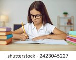 Portrait of clever school child. Little student girl in glasses sitting at desk, studying new things and writing in notebook. Learning and education concept