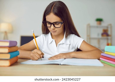 Portrait of clever school child. Little student girl in glasses sitting at desk, studying new things and writing in notebook. Learning and education concept