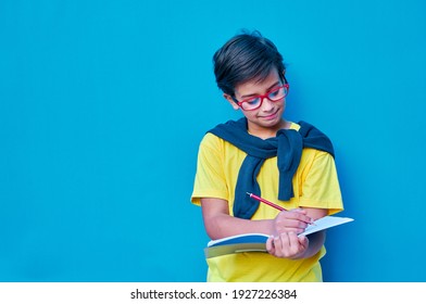 A Portrait of a clever and studious boy with red glasses, with a yellow shirt and a sweater on his shoulders, holding a pencil and a notebook in his hand to study. On blue background. Copy space