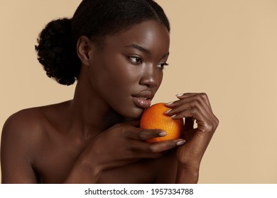 Portrait close up of beautiful african girl hold whole of orange. Serious young woman with perfect skin. Concept of skincare. Isolated on beige background. Studio shoot