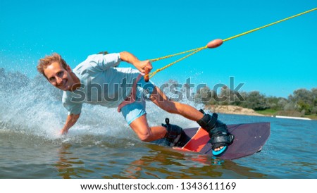 PORTRAIT, CLOSE UP Cheerful young surfer wakeboarding, sliding his hand on water surface, splashing water at camera. Smiling wakesurfer riding waterski cable park on sunny day. Wakesurfing in summer
